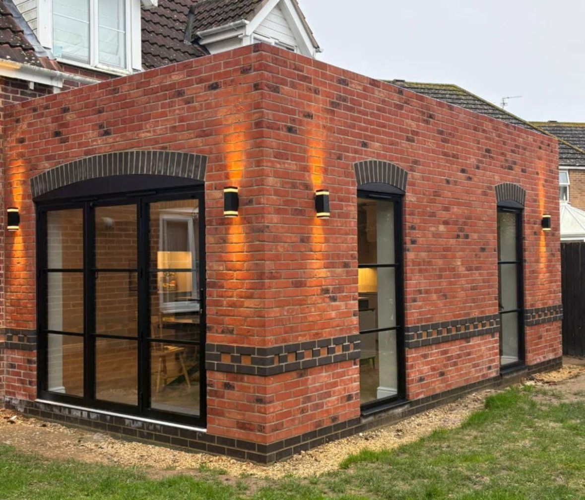 Red-brick flat-roof extension with arched black steel-framed doors and wall lights