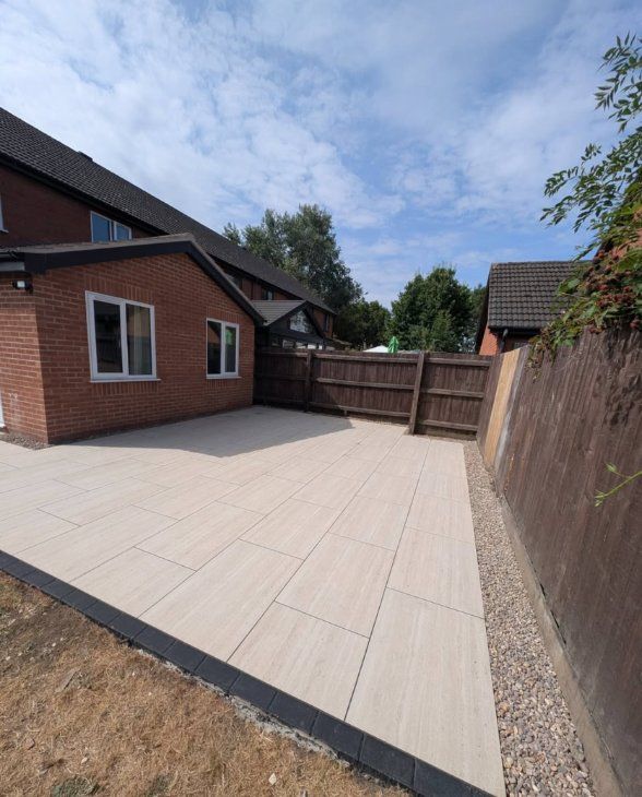 Large light-grey porcelain patio with dark edging and a gravel border beside a red-brick house